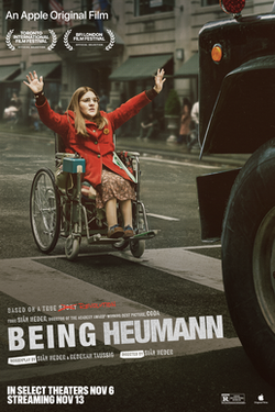 Movie poster for Being Heumann. A woman portraying Judy Heumann sits in a wheelchair in the middle of a city street, arms raised high, wearing a red coat and glasses. The front of a large black vintage vehicle looms at the right edge.