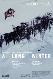Movie poster for A Long Winter. Seen from above, a lone figure in a dark coat stands in deep snow at the top left. Below, dark pine trees line a white, snow-covered slope. The title appears in widely spaced white capitals. January 22.