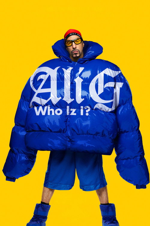 Movie poster for Ali G: Who Iz I? A man in sunglasses and a red cap stands against a bright yellow background, almost swallowed by an enormous blue puffer jacket, with matching blue shorts and boots. The title is printed across the jacket in white letters.