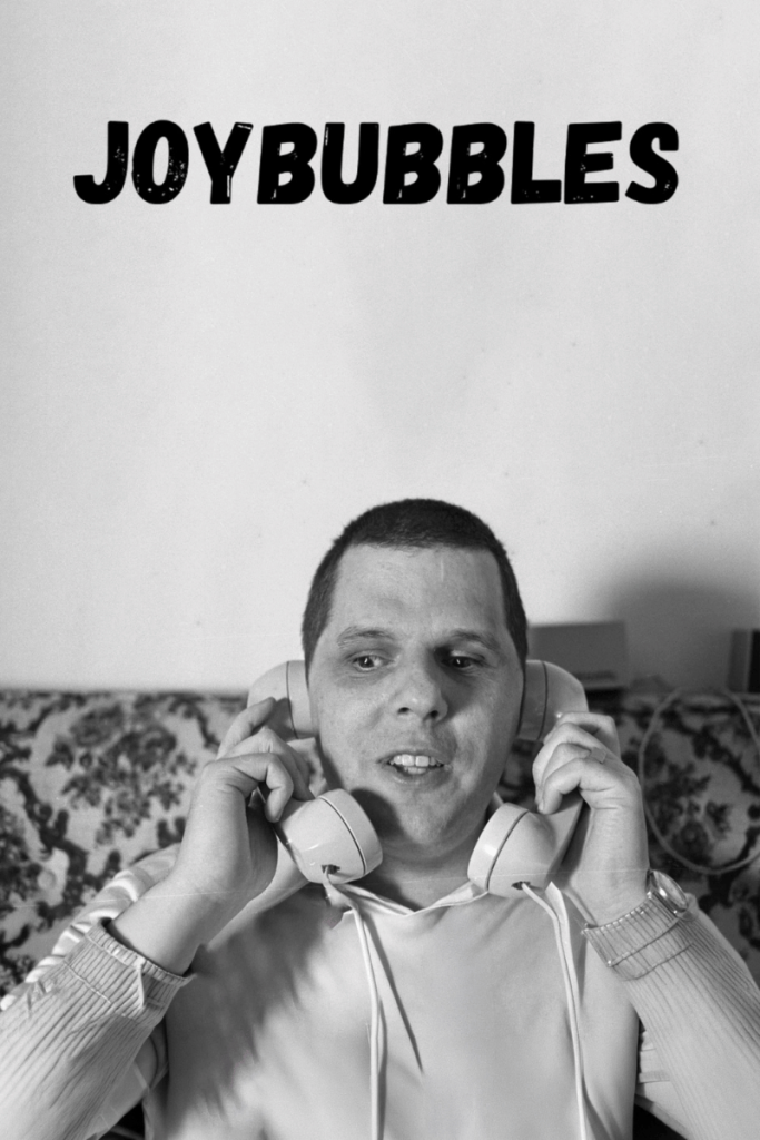 Movie poster for Joybubbles. A black and white photograph of a man sitting on a patterned sofa, holding two telephone handsets up to his ears, mouth open mid-speech. The wall behind him is plain and empty. The title appears across the top in black capitals.