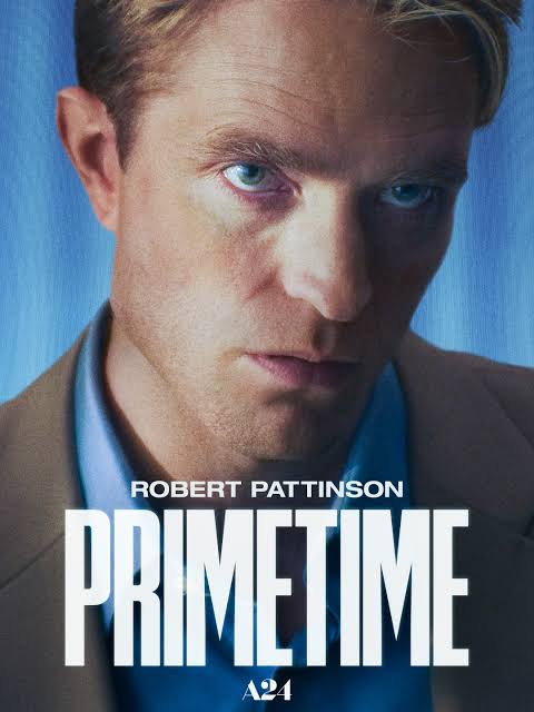 Movie poster for Primetime. Extreme close up of a man's face filling the frame, tilted slightly and lit in cold blue, with a distorted second eye visible on the left side. He wears a tan jacket over a blue collared shirt. The title appears at the bottom in large white capitals, with the name Robert Pattinson above it.