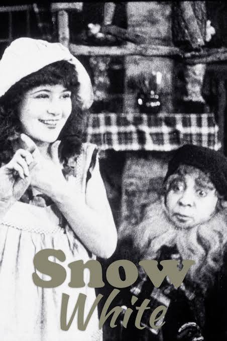 Movie poster for Snow White. A black and white still shows a young woman in a white bonnet and pinafore smiling toward the camera in a rustic cabin interior. Beside her stands a shorter figure in a dark cap and heavy fur-trimmed coat, looking up with a startled expression. The title appears across the bottom in pale green lettering.